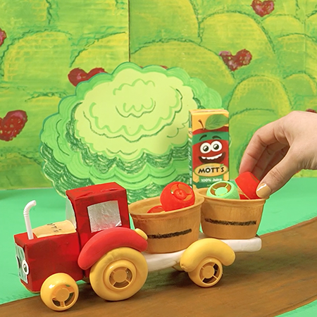Hand placing an apple in a basket sitting on the back of a  DIY red farm truck.  Motts apple juice box in the background.