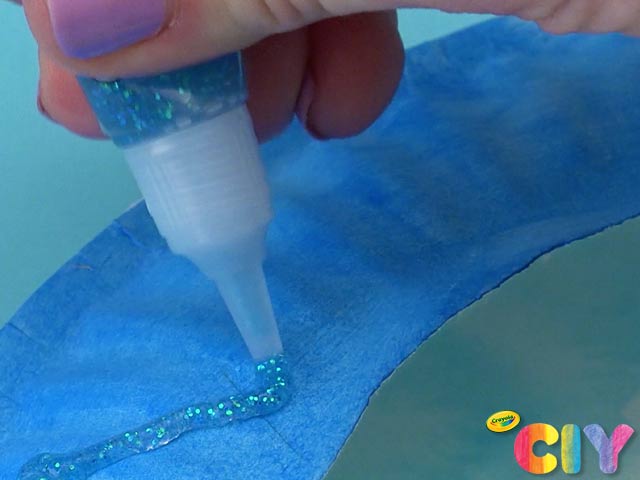 Hand squeezing Crayola Glitter Glue onto blue-painted edge of paper plate