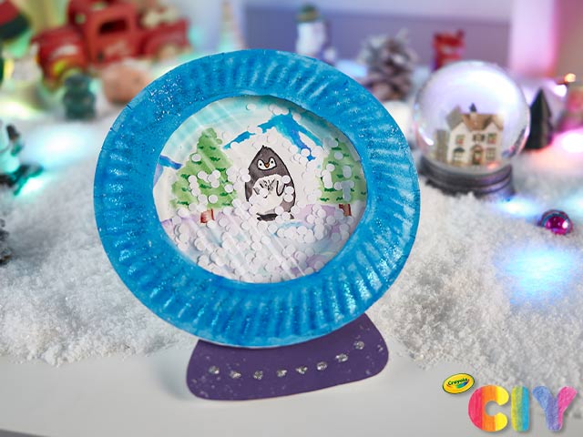 Homemade paper plate snow globe standing upright with fake snow and winter decorations on table