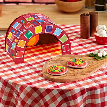 Cardboard Pizza Oven Craft sitting on a red checker tablecloth with Model Magic Pizzas