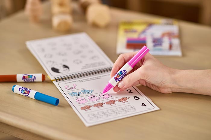 Color & Erase Reusable Activity Pad, Numbers. Hand holding color & erase marker over open activity pad on table.